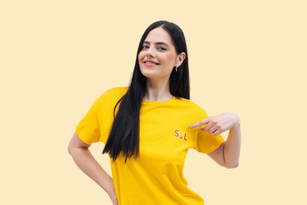 Woman with long dark hair wearing a yellow t-shirt that says "SOL," pointing at the shirt and smiling, standing against a solid light yellow background.